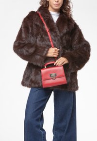 Manteau en fausse fourrure marron avec un large col, associé à un sac à main en cuir rouge avec une fermeture dorée et une poignée supérieure. Le mannequin porte un jean bleu.