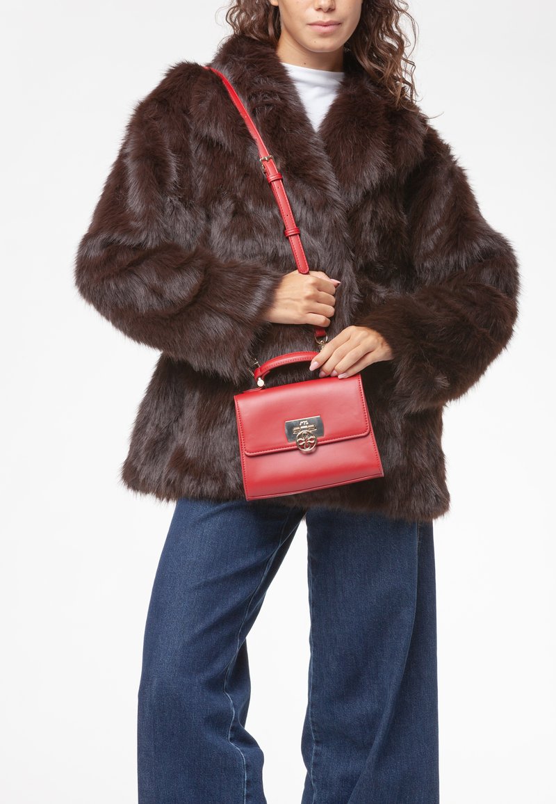 Manteau en fausse fourrure marron avec un large col, associé à un sac à main en cuir rouge avec une fermeture dorée et une poignée supérieure. Le mannequin porte un jean bleu.