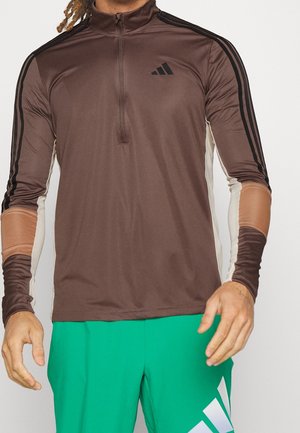 Mężczyzna w brązowej, długorękawowej bluzie Adidas z czarnymi paskami i zielonych spodenkach Adidas z białym logo, stojący na białym tle.