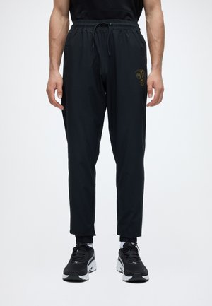 Hombre con pantalones negros de cintura elástica con un sutil logo dorado, combinados con zapatillas deportivas en blanco y negro y calcetines blancos.
