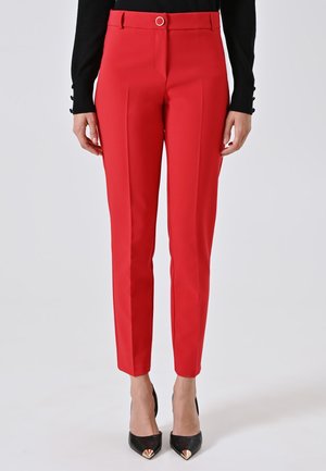 Pantaloni rossi slim-fit con pieghe frontali, vita con bottone argentato, indossati con una maglietta nera a maniche lunghe e tacchi a punta neri.