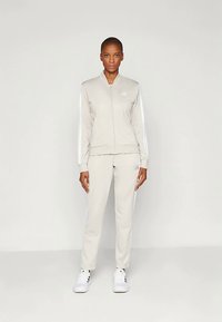 Ανοιχτό γκρι αθλητικό σύνολο που περιλαμβάνει μια ζακέτα bomber με φερμουάρ και ταιριαστά παντελόνια. Λευκές ρίγες τονίζουν τους ώμους και τις πλευρές. Λευκά αθλητικά παπούτσια ολοκληρώνουν την εμφάνιση.