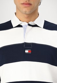 Marineblå og hvit stripete polo skjorte med krage og knapper, med horisontalt stripedesign og "TOMMY JEANS"-logo på brystet.