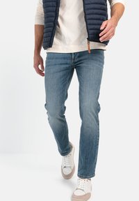Lyseblå denimjeans med lige pasform, parret med en cremefarvet langærmet skjorte og en marineblå puffervest. Hvide sneakers med beige detaljer.