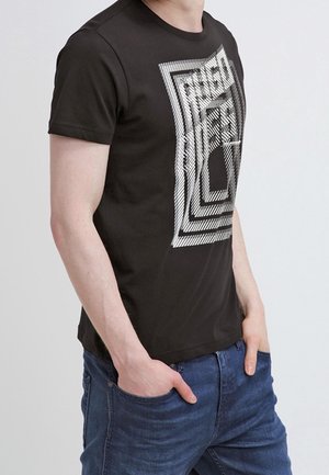 Schwarzes kurzärmeliges T-Shirt mit weißem geometrischem Quadratmuster und abstraktem Text, getragen von einer Person mit Händen in den Taschen einer blauen Jeans.