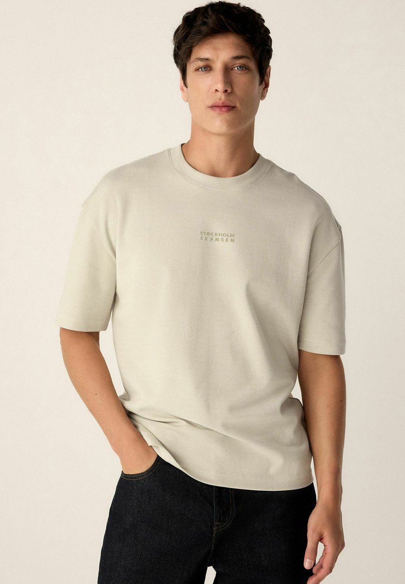 Camiseta de manga corta de color beige claro con una textura acanalada y un logo sutil centrado en el pecho. Combinada con jeans de mezclilla oscura.