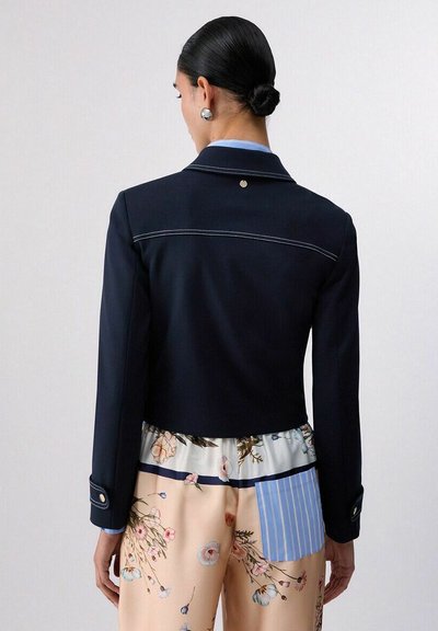 Femme aux cheveux noirs en chignon, portant un blazer court bleu marine et un pantalon large à fleurs avec une poche arrière rayée bleue, posant devant un fond uni.