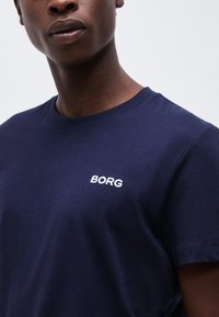 Νεαρό άτομο που φοράει μπλε σκούρο μπλουζάκι με το κέντημα «BORG» σε λευκό στην αριστερή πλευρά του στήθους, με φόντο ένα απλό σκηνικό.