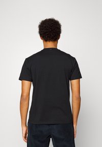 Hombre con cabello rizado, vistiendo una camiseta negra de manga corta lisa y pantalones oscuros, de pie de espaldas contra un fondo claro.