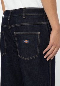 Dark blue denim jeans featuring yellow stitching, a logo patch on the back pocket, and a fitted design with a classic five-pocket style.