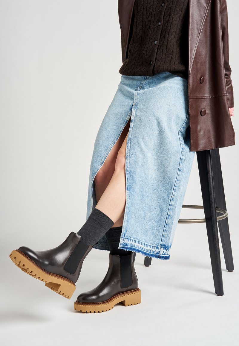 Botas de tobillo de cuero negro con una suela gruesa marrón, combinadas con una falda de mezclilla azul clara que presenta una abertura lateral y calcetines gris oscuro.