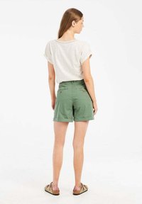 Lichtbeige shirt met opgerolde mouwen, gecombineerd met groene shorts met zakken en knopen, gedragen met bruine slipper sandalen.