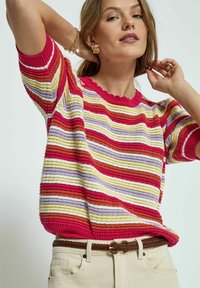 Gestreifter Pullover in Rot, Pink, Gelb und Lavendel, mit einem gerippten Textur und einem gewellten Ausschnitt. Kombiniert mit beigen Hosen und einem geflochtenen Gürtel.