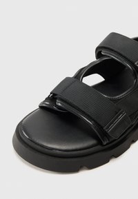 Black sandal featuring adjustable fabric straps, a cushioned footbed, and a thick, rugged rubber sole. Smooth leather finish on the upper.