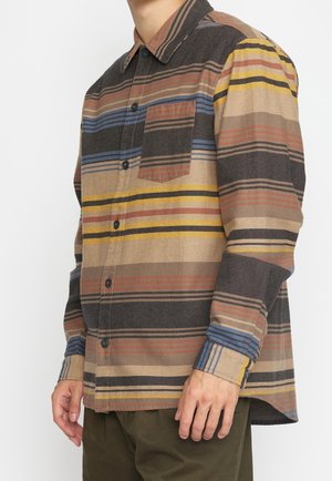 Mężczyzna w koszuli na długi rękaw z guzikami, z poziomymi paskami w odcieniach brązu, żółci, niebieskiego i czerni oraz z kieszenią na piersi po lewej stronie.
