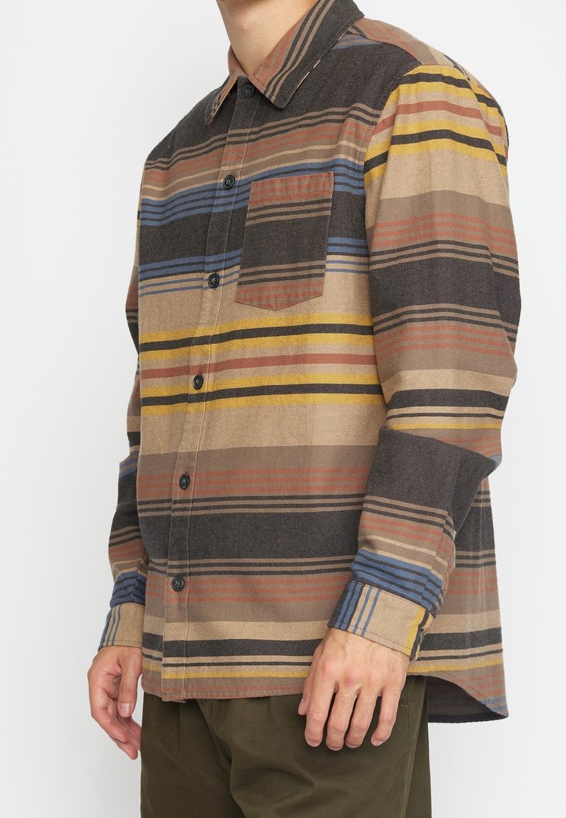 Homme portant une chemise à manches longues boutonnée avec des rayures horizontales de couleurs marron, jaune, bleu et noir, avec une poche sur la poitrine côté gauche.