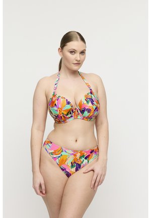 Vrouw met lichte huid en bruin haar in een paardenstaart, gekleed in een kleurrijke bloemenhalterbikini, poseert tegen een witte achtergrond.