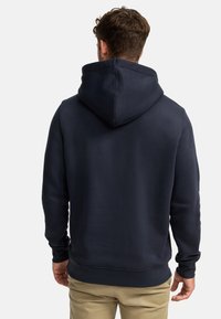 Bărbat purtând un hanorac cu glugă de culoare bleumarin închis și pantaloni bej, întors cu spatele, pe un fundal simplu, deschis la culoare.