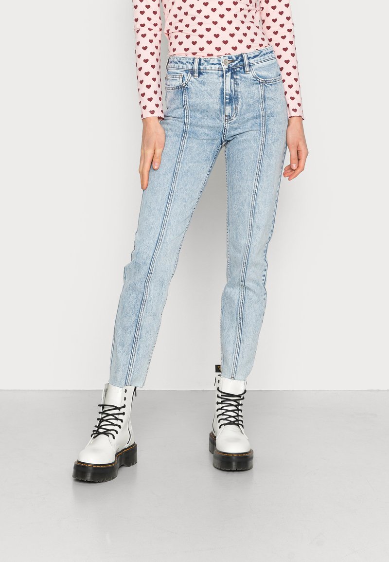 Jeans de cintura alta azul claro com costuras verticais, emparelhados com botas brancas de atar. O material tem uma textura de denim ligeiramente desbotada.