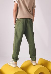 Grüne Cargo-Hosen mit Seitentaschen, aus leichtem Stoff, kombiniert mit beigefarbenen Sneakers. Das Oberteil ist beige mit hochgerollten Ärmeln.