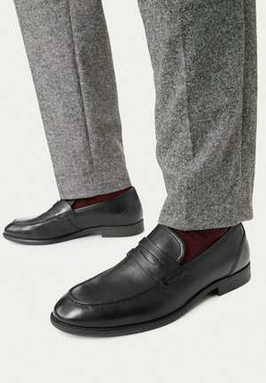 Schwarze Lackleder-Herrenschuhe zum Hineinschlüpfen, getragen mit grauen Hosen und dunkelroten Socken, dargestellt auf hellem Hintergrund.