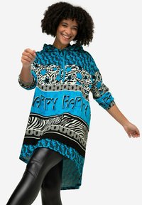 Gewatteerde blauwe tuniek met dierenprints en de tekst "Happy." Beschikt over zwart-witte zebraprinten en gedurfde patronen; relaxed fit en lange mouwen.