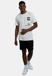 Camiseta gris claro con logo y texto en negro, combinada con pantalones cortos negros. Los accesorios incluyen zapatillas blancas, calcetines de tobillo a juego y una pose casual.