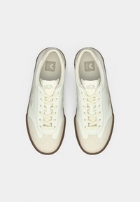 Bílé kožené tenisky s kulatou špičkou, béžovými akcenty a šesti očními dírkami. Texturované detaily na špičce a bocích. Hnědá gumová podrážka.