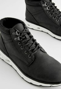 Czarne skórzane botki do kostki z białymi gumowymi podeszwami, z metalowymi oczkami i czarnymi sznurowadłami. Teksturowana powierzchnia i wyściełany kołnierz.