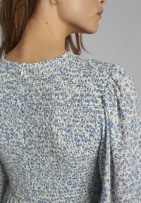 Sukienka w kwiaty z marszczeniem w odcieniu kremowym z niebieskimi wzorami. Posiada bufiaste rękawy oraz zapięcie na guziki z tyłu. Szczegóły z teksturowanej tkaniny.