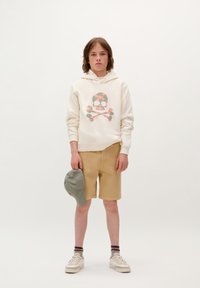 Lichtbeige hoodie met een bloemen schedel graphic, tan shorts, grijze pet en crèmekleurige sneakers. De look is casual en ontspannen.