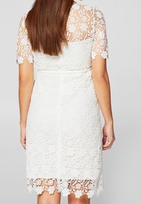 Vit spetsklänning med korta ärmar, med ett blommigt mönster och vågkant. Stoffet har en texturerad, ömtålig utseende.