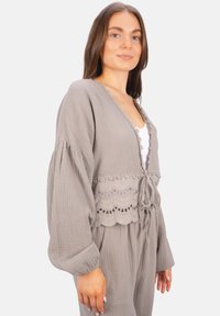 Grau gefärbter Stoffmantel mit voluminösen Ärmeln, Spitzendetails und Bindeband in der Taille. Leichter Stoff mit einem gewellten Muster am Saum.