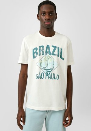 Young man wearing white t-shirt with faded "Brazil São Paulo" text and palm tree graphic, paired with light blue jeans.
