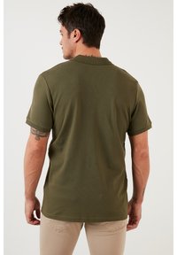 Camisa de manga corta de canalé verde oliva con cuello redondeado, detalles de costura visibles y un corte relajado, vista desde la espalda de un modelo.