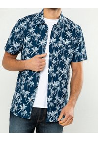 Camicia a maniche corte di colore blu navy con motivo di palme bianche, con bottone frontale, tessuto testurizzato, abbinata a una maglietta bianca e jeans blu.