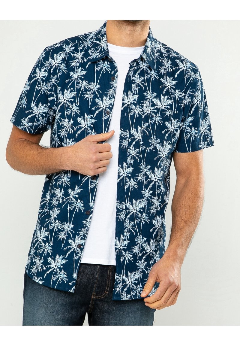 Camicia a maniche corte di colore blu navy con motivo di palme bianche, con bottone frontale, tessuto testurizzato, abbinata a una maglietta bianca e jeans blu.