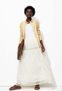 Jupe maxi blanche avec des motifs en œillets, associée à une chemise à rayures jaune clair et un sac à main en franges marron. Des sandales à lanières marron complètent le look.