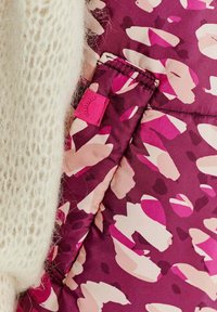 Prošívaná látka v odstínech fialové s abstraktními růžovými a krémovými vzory. Obsahuje nadýchanou bílou pletenou texturu a malou růžovou štítkovou dekoraci.