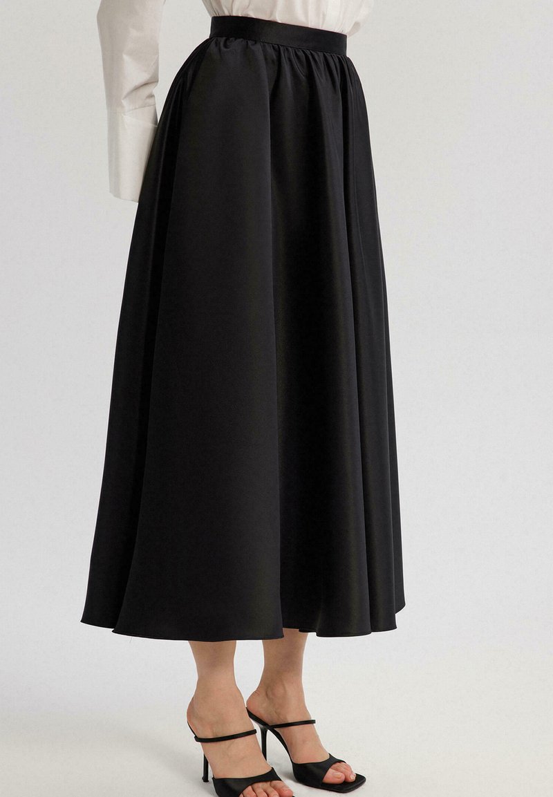 Jupe maxi noire en tissu lisse, avec une taille froncée et un design fluide, associée à des talons à lanières noirs.
