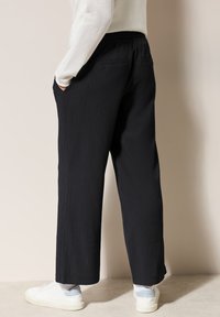 Pantalones de pierna ancha negros y texturizados con bolsillos laterales y un corte relajado. Combinados con zapatillas blancas y calcetines claros en contraste.