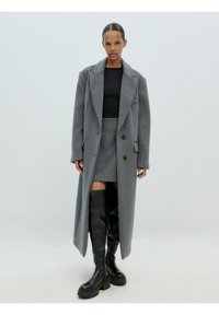 Un long manteau gris à rayures avec un design ajusté, associé à une jupe assortie. Des bottes noires montantes avec une semelle épaisse.