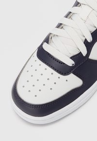 Tenis cu vârf alb și bleumarin închis, cu găuri perforate și șireturi albe, pe un fundal deschis.
