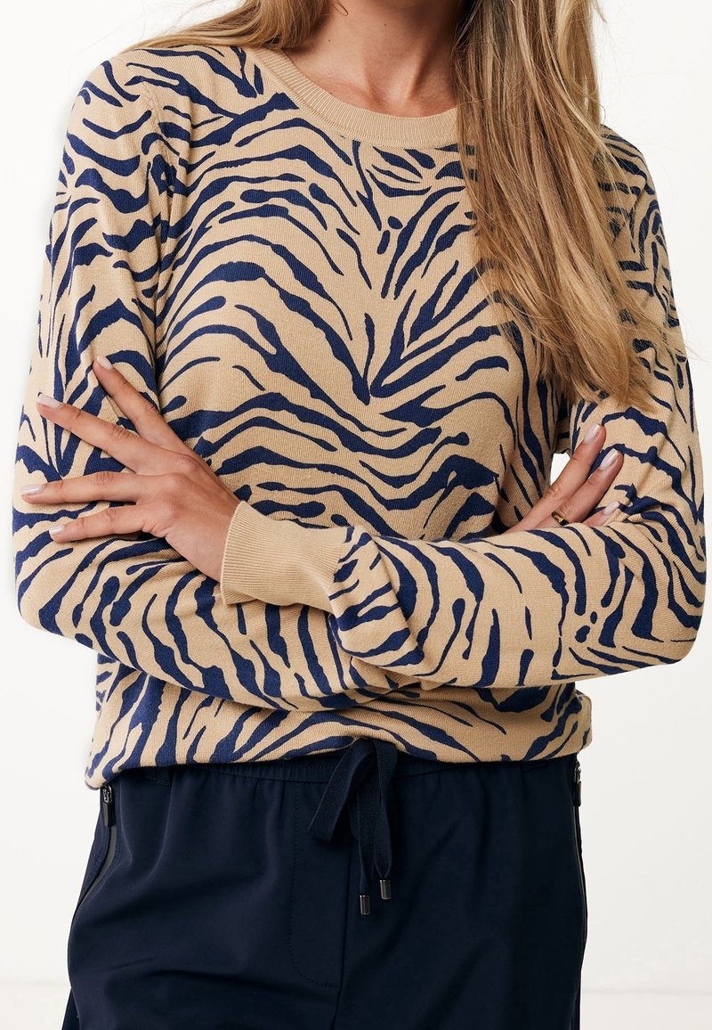 Femme aux longs cheveux portant un pull à imprimé zèbre beige et bleu marine, les bras croisés sur un pantalon bleu marine à taille élastiquée avec cordon.