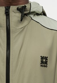 Μπεζ ζακέτα με κουκούλα, με μαύρο φερμουάρ, μαύρη κορδόνι και μαύρο λουλουδάτο λογότυπο Hugo στο στήθος.