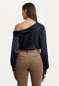 Navy satijnen off-shoulder blouse met lange mouwen, voorzien van een gedrapeerde kraag. Kombineert met beige corduroy broek en een bruine riem.