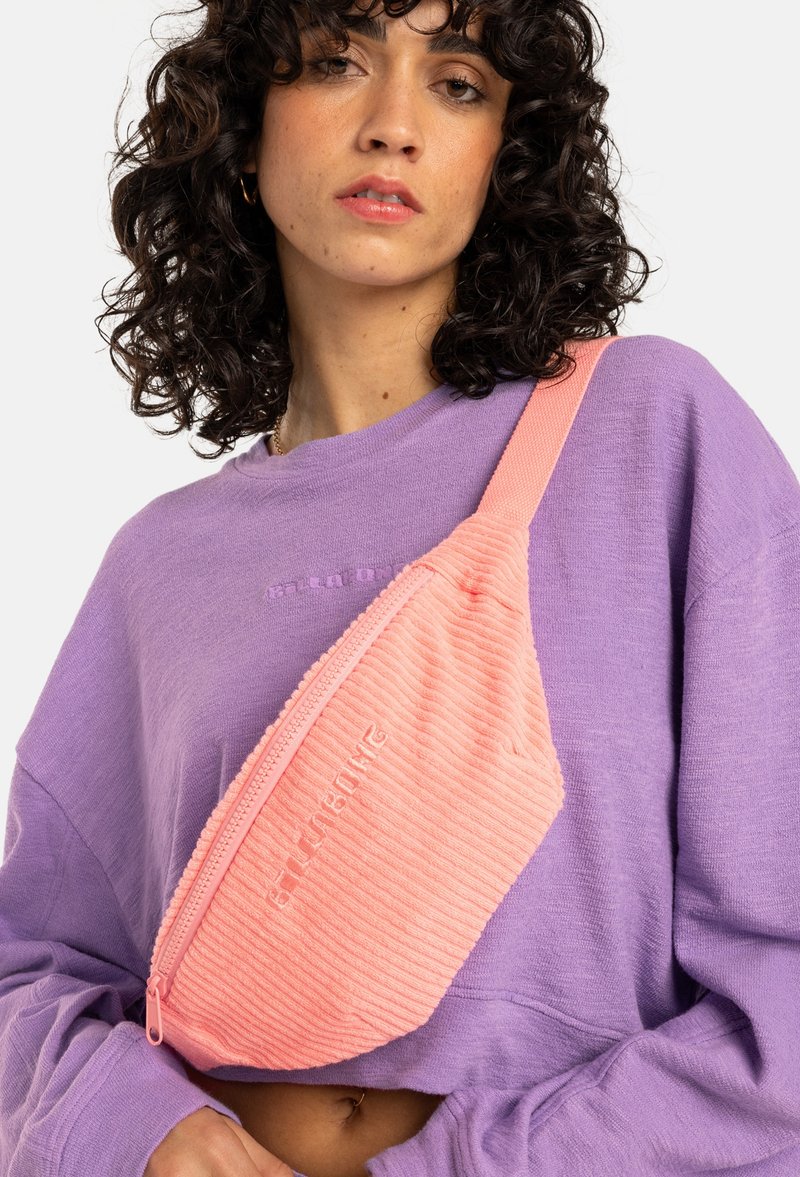 Korallrosa kordflöjtsaxelväska med dragkedjestängning och präglad logotyp. Bärs över en lavendelfärgad oversized sweatshirt. Slät textur.
