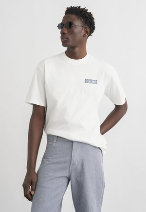 Hombre con gafas de sol redondas negras, camiseta blanca de Dickies y pantalones de rayas azules y blancas, de pie con una mano detrás de la espalda.