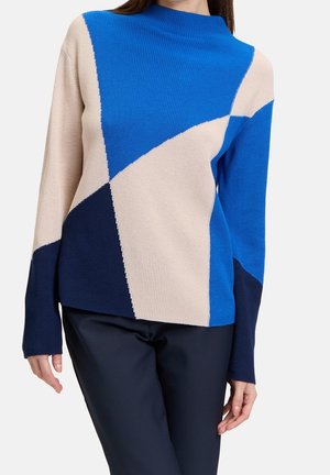 Kvinde iført en langærmet, farveblok-strikketrøje i blå, beige og marineblå, kombineret med mørke marineblå bukser mod en ensfarvet baggrund.