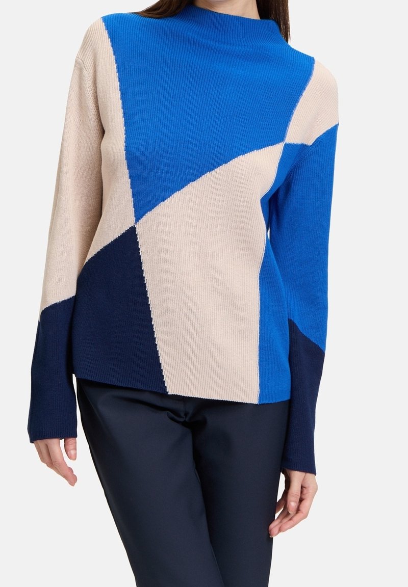 Femme portant un pull en maille à manches longues et blocs de couleurs bleu, beige et marine, associé à un pantalon bleu marine foncé sur un fond uni.